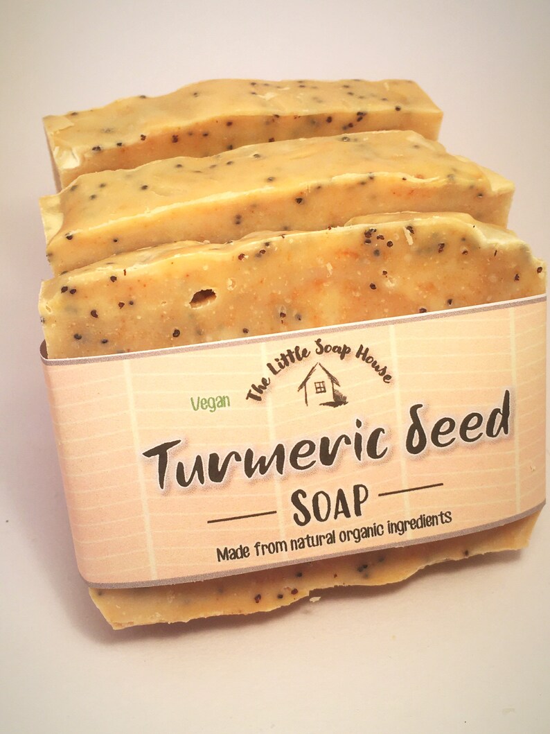 K&ouml;nnte beinhalten: Drei Seifenst&uuml;cke mit Kurkuma-Samen, hellorangefarben mit dunklen Samenflecken. Die Seife ist mit einem Etikett versehen, auf dem "Turmeric Seed Soap" und "Made from natural organic ingredients" mit dem Logo "The Little Soap House" steht.