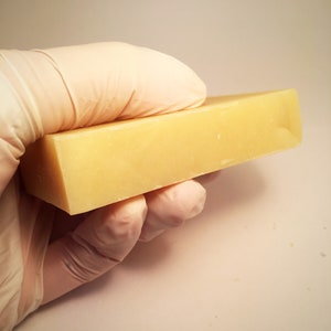 May include: A rectangular bar of pale yellow soap, held in a gloved hand.