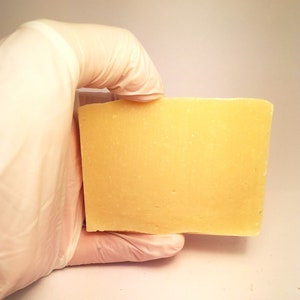 May include: A rectangular bar of pale yellow soap, held in a gloved hand.