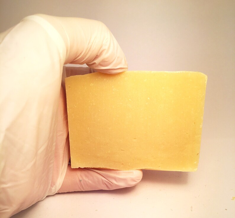 May include: A rectangular bar of pale yellow soap, held in a gloved hand.