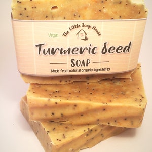 K&ouml;nnte beinhalten: Gestapelte St&uuml;cke Kurkuma-Samen-Seife. Die Seife hat eine hellgelbe Farbe mit dunklen Punkten. Ein Papieretikett umschlie&szlig;t das oberste St&uuml;ck, auf dem die Worte "Turmeric Seed Soap" aufgedruckt sind. Das Etikett enth&auml;lt auch die Worte "Vegan" und "Made from natural organic ingredients."
