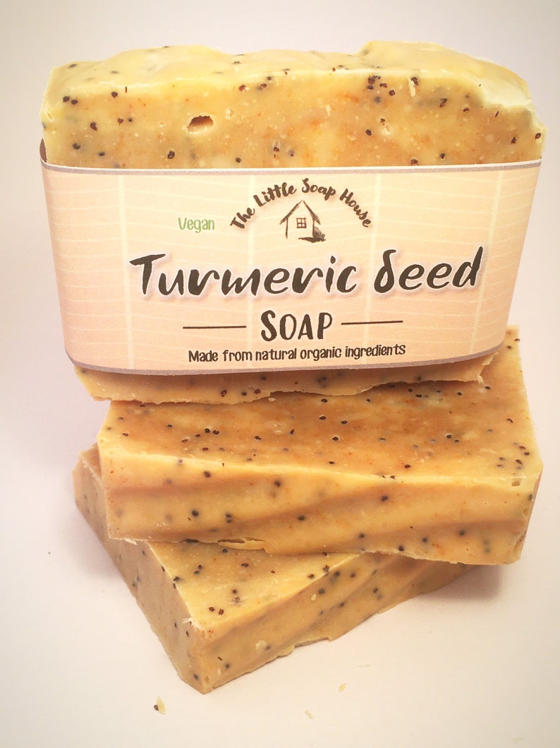 K&ouml;nnte beinhalten: Gestapelte St&uuml;cke Kurkuma-Samen-Seife. Die Seife hat eine hellgelbe Farbe mit dunklen Punkten. Ein Papieretikett umschlie&szlig;t das oberste St&uuml;ck, auf dem die Worte "Turmeric Seed Soap" aufgedruckt sind. Das Etikett enth&auml;lt auch die Worte "Vegan" und "Made from natural organic ingredients."