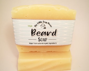 May include: A rectangular block of pale yellow beard soap wrapped in a light blue paper label. The label features the text The Little Soap House, Vegan, Beard SOAP, and Made from natural organic ingredients with a small house icon illustration.