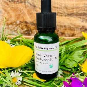 Puede incluir: Una botella de vidrio verde con un gotero negro, etiquetada "The Little Soap House Aloe Vera & Hyaluronic Acid Serum 1.5%". La botella est&aacute; rodeada de hierba verde, flores amarillas y blancas.