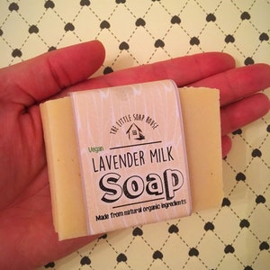 May include: A bar of lavender milk soap in a hand. The soap is wrapped in a white label with the text "The Little Soap House", "Vegan", "Lavender Milk Soap", and "Made from natural organic ingredients".