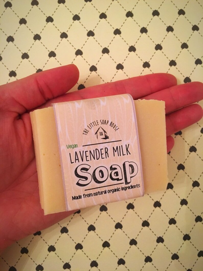 May include: A bar of lavender milk soap in a hand. The soap is wrapped in a white label with the text "The Little Soap House", "Vegan", "Lavender Milk Soap", and "Made from natural organic ingredients".