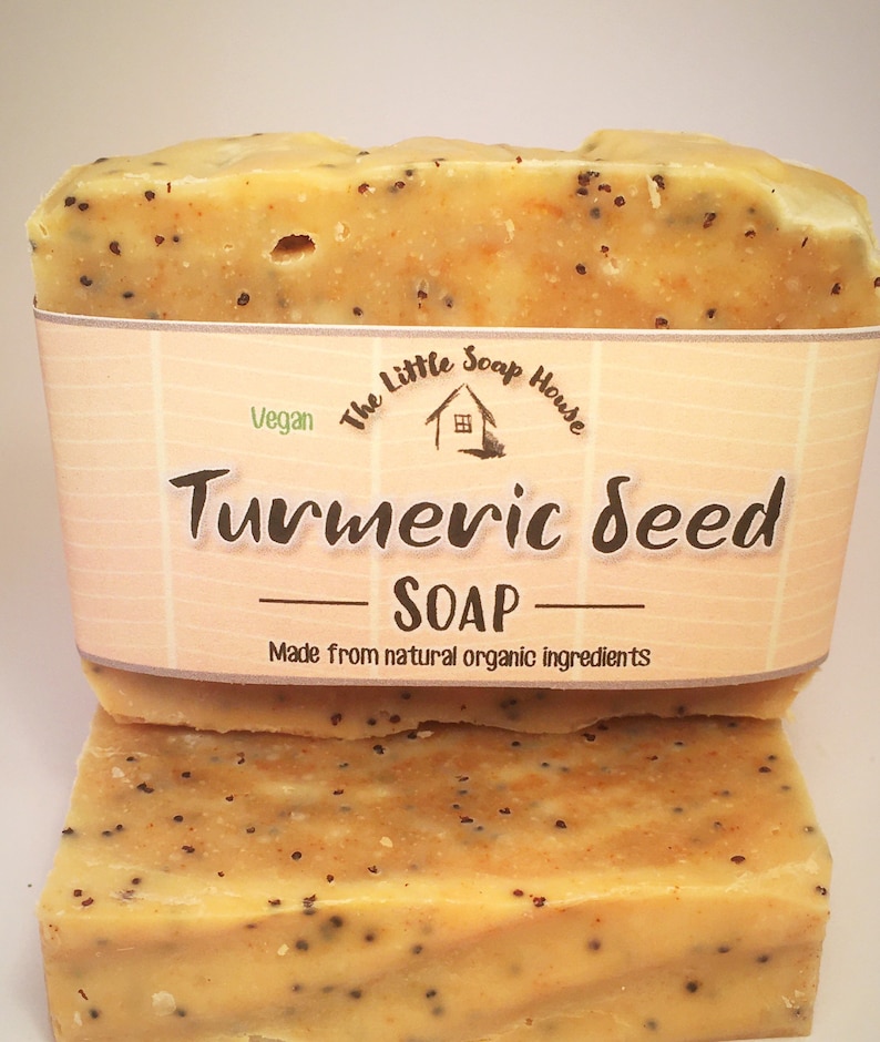 K&ouml;nnte beinhalten: Zwei Seifenst&uuml;cke mit Kurkuma-Samen, hellorangefarben mit dunklen Flecken. Die Seife ist mit einem Etikett versehen, auf dem "Turmeric Seed Soap" und "Made from natural organic ingredients" steht. Das Etikett zeigt auch ein kleines Hauslogo.