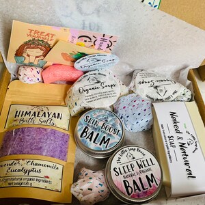 May include: A gift box filled with bath and body products, including bath salts, soap, and balms. The box is lined with white tissue paper and has a brown cardboard exterior. The products are labeled with the text "Himalayan Bath Salts", "Skin Boost Balm", "Sleep Well Balm", and "Naked & Natural Soap".