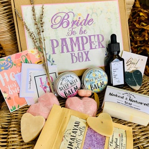 Op de afbeelding: Een rieten mand gevuld met verschillende verzorgingsproducten, waaronder badzout, zeep en balsems. De mand is gelabeld met "Bride to be Pamper Box".