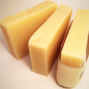 May include: Three bars of pale yellow soap, two are stacked on top of each other, and the third is wrapped in brown paper with a white label.