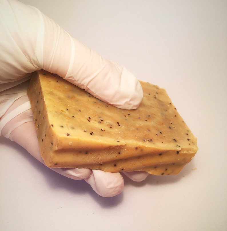 K&ouml;nnte beinhalten: Ein rechteckiger Block handgemachter Seife mit hellbrauner Farbe und kleinen schwarzen Punkten. Die Seife hat eine strukturierte Oberfl&auml;che und wird von einer behandschuhten Hand gehalten. Die Seife scheint frisch geschnitten zu sein, mit sichtbaren Rillen auf einer Seite.
