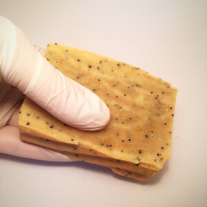 K&ouml;nnte beinhalten: Drei rechteckige St&uuml;cke handgemachte Seife in hellem Beige mit kleinen schwarzen Punkten. Eine behandschuhte Hand h&auml;lt die Seife vor einem wei&szlig;en Hintergrund. Die Seife scheint gestapelt zu sein.