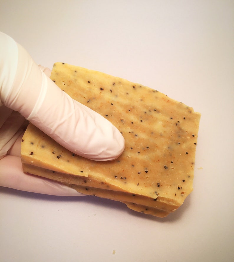 K&ouml;nnte beinhalten: Drei rechteckige St&uuml;cke handgemachte Seife in hellem Beige mit kleinen schwarzen Punkten. Eine behandschuhte Hand h&auml;lt die Seife vor einem wei&szlig;en Hintergrund. Die Seife scheint gestapelt zu sein.