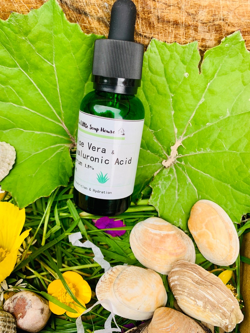 Puede incluir: Una botella de vidrio verde con un gotero negro. La botella est&aacute; etiquetada "Aloe Vera & &Aacute;cido Hialur&oacute;nico Suero 1.5%" y tiene una peque&ntilde;a ilustraci&oacute;n de planta de aloe vera. La botella est&aacute; sentada en una cama de hierba verde y conchas.