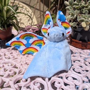 May include: A soft, light blue bat toy with rainbow detailing on the wings and ears. The wings display a blue sky design with rainbows. The bat is on a pink table, with plants in the background.