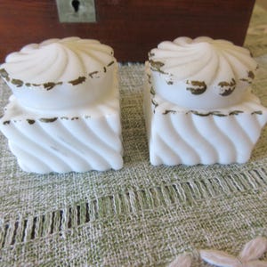 May include: Two white, antique, square-shaped salt cellars with a swirled design. The lids have a spiral pattern and are trimmed with a gold-colored accent. The cellars sit on a green and white patterned surface.