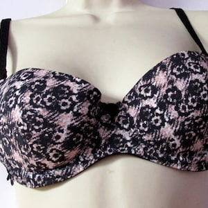 May include: A strapless-style bra featuring a black and light pink floral pattern across the cups. The garment includes thin, adjustable black shoulder straps and a small black bow detail centered between the two molded cups.