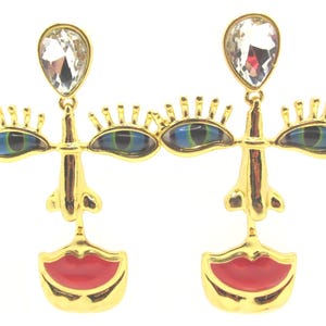 Peut inclure: Une paire de boucles d'oreilles dorées en forme de visages abstraits. Chaque boucle présente un cristal transparent en forme de goutte en haut, deux accents en forme d'yeux bleus et verts avec des cils dorés, et des lèvres en émail rouge.