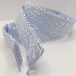 Pode incluir: Fita azul clara com um padrão floral e geométrico. A fita está dobrada sobre si mesma, mostrando os detalhes intrincados do design. As bordas são finalizadas com uma borda de contas.