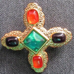 May include: A gold-toned cross-shaped brooch with four colorful stones. The center stone is a large, emerald green square. The other three stones are smaller, oval-shaped, and red.