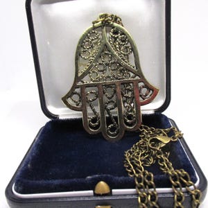 May include: A gold-coloured Hamsa hand pendant necklace with an elaborate openwork design. The pendant is displayed in an open, black jewellery box with a blue velvet interior. The necklace chain is also gold-coloured.