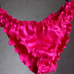 May include: Bright pink satin underwear with ruffled edges. The lingerie has a shiny, smooth texture and a delicate, feminine design. The elastic waistband is gathered, and the leg openings are trimmed with ruffles.
