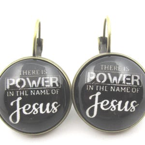 Könnte beinhalten: Ein Paar runde Ohrringe mit schwarzem Hintergrund und dem Text "THERE IS POWER IN THE NAME OF Jesus" in Weiß. Die Ohrringe haben einen bronzefarbenen Metallrahmen und Ohrhaken.