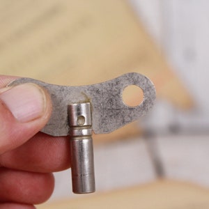 Può includere: Piccola chiave metallica vintage con corpo cilindrico e testa piatta e irregolare. La testa presenta un foro circolare e una piccola apertura rettangolare. La chiave è tenuta in mano, suggerendo il suo utilizzo per caricare un dispositivo meccanico.