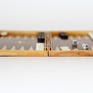May include: A wooden backgammon board with a brown playing surface and cream and black game pieces. Two dice are visible on the board. The board is open and ready for play.