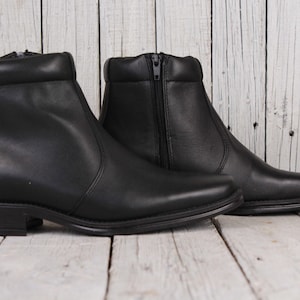 Peut inclure: Paire de bottines en cuir noir avec un petit talon. Les bottines ont une fermeture &eacute;clair sur le c&ocirc;t&eacute; et un bout arrondi. Elles sont pr&eacute;sent&eacute;es sur une surface en bois clair.