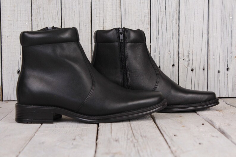 Peut inclure: Paire de bottines en cuir noir avec un petit talon. Les bottines ont une fermeture &eacute;clair sur le c&ocirc;t&eacute; et un bout arrondi. Elles sont pr&eacute;sent&eacute;es sur une surface en bois clair.