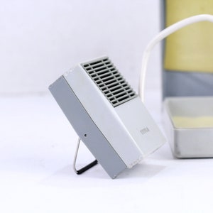 May include: A white and gray small electric heater with a metal grill on the front. The heater is on a small metal stand and has a cord attached to it. The heater is sitting on a white surface.