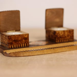 Puede incluir: Un tintero de madera con dos compartimentos con bisagras, cada uno con un pequeño pozo de porcelana blanca. El tintero está decorado con intrincadas tallas de madera y se asienta sobre una base de madera.