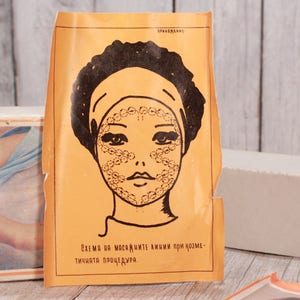May include: Vintage orange paper with a black illustration of a woman's face, featuring a headband and dotted patterns on the face. Text in Russian is visible on the paper and a box in the background. The image suggests a skincare or beauty product.