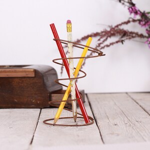 May include: A decorative pencil holder made of brown metal wire, holding three pencils. One pencil is red, one is yellow, and one is white with a floral pattern. The holder has a spiral design and sits on a white wooden surface.