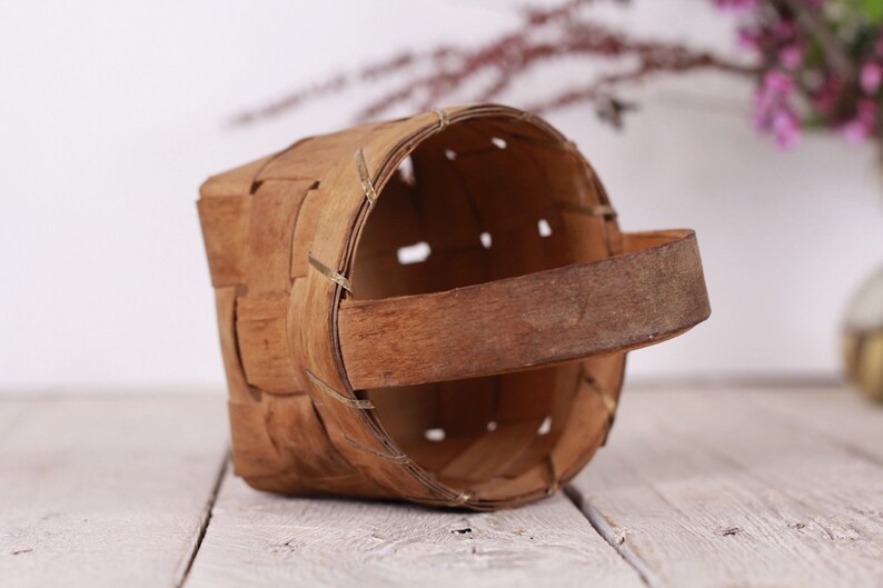 Puede incluir: Una peque&ntilde;a cesta artesanal hecha de tiras de madera tejidas. La cesta tiene forma rectangular con la parte superior redondeada y un asa resistente. Los tonos naturales de la madera y el dise&ntilde;o sencillo le dan un aspecto r&uacute;stico y vintage.
