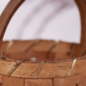 Puede incluir: Una peque&ntilde;a cesta de madera hecha a mano con un asa curva. La cesta est&aacute; hecha de madera de color marr&oacute;n claro y presenta detalles met&aacute;licos decorativos alrededor del borde. El asa tambi&eacute;n es de madera.