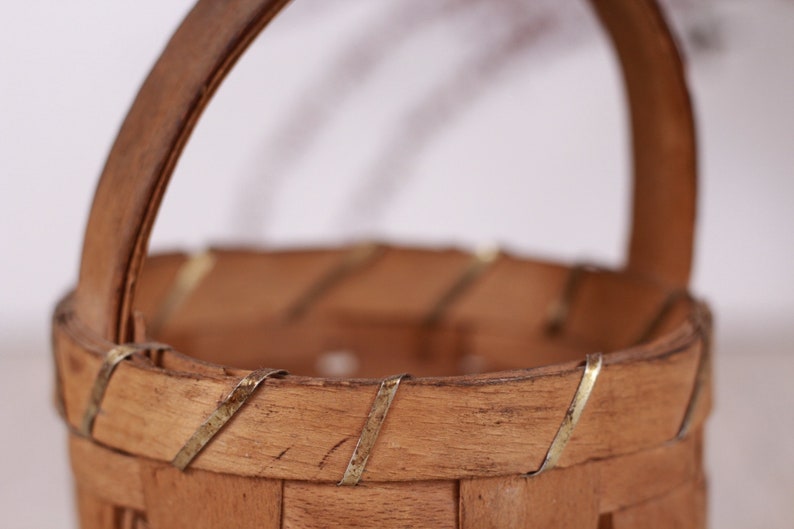 Puede incluir: Una peque&ntilde;a cesta de madera hecha a mano con un asa curva. La cesta est&aacute; hecha de madera de color marr&oacute;n claro y presenta detalles met&aacute;licos decorativos alrededor del borde. El asa tambi&eacute;n es de madera.