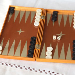 May include: A folding backgammon board with an orange plastic frame and a brown playing surface featuring gold triangular points. The set includes black and white circular checkers arranged on the board and two small white dice with black pips.