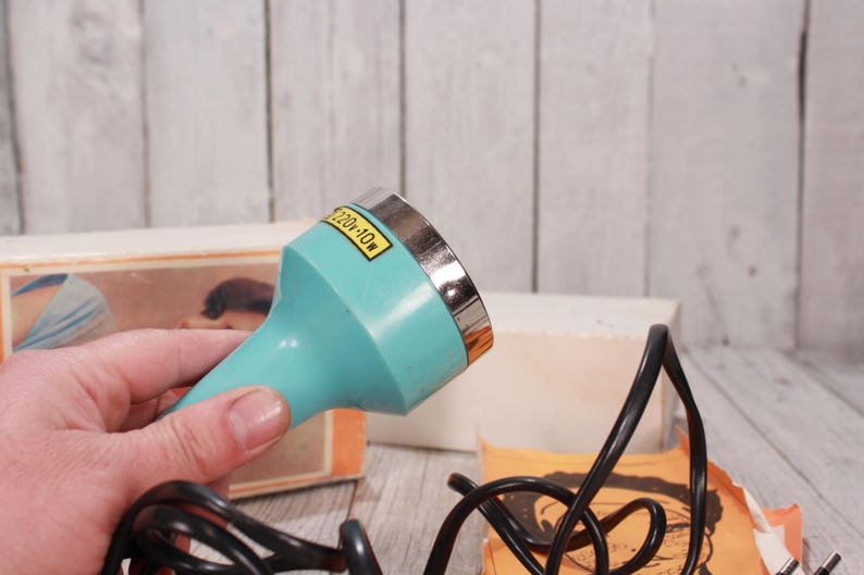 May include: A vintage teal-colored appliance with a silver rim and a black power cord. The appliance has a label that reads "220v-10w". The background includes a box with an image and a wooden surface.