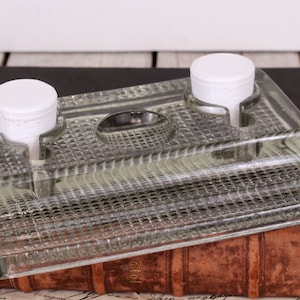 May include: A clear glass inkwell with two white-topped ink bottle holders and a central pen rest. The inkwell has a textured, diamond-patterned surface and sits on a dark surface, resting on a vintage book.