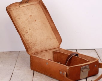 May include: A brown leather camera case with a strap and a metal latch. The case is open and the inside is lined with brown fabric.