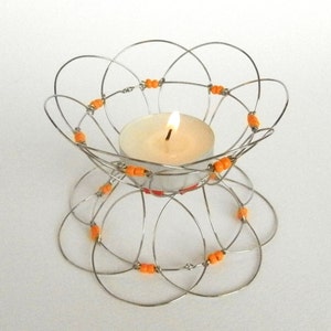 May include: A silver wire candle holder with a floral design, adorned with orange beads. A lit, white tea light candle sits in the center. The candle holder is on a white surface.
