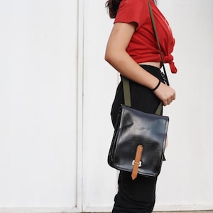 May include: A black leather crossbody bag with a brown strap and a green strap. The bag is worn over the shoulder of a person wearing a red shirt and black pants.