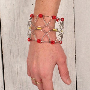 May include: A silver-toned wire bracelet with red beads and yellow accents. The bracelet is worn on a wrist, showcasing its intricate design and the contrast between the silver, red, and yellow elements. The bracelet's design is geometric and eye-catching.