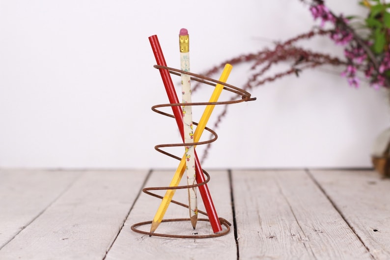 May include: A rustic, spiral-shaped pencil holder made of brown metal, holding three pencils. One pencil is red, one is yellow, and the third is white with a floral pattern. The pencils are arranged in a decorative display.