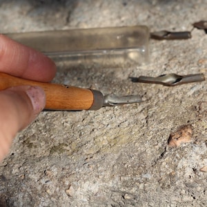 Könnte beinhalten: Ein Holz-Federhalter mit einer Metallfeder, gehalten von einer Hand. Mehrere Metallfedern und eine transparente Kunststoffhülle sind ebenfalls auf einer strukturierten, grauen Oberfläche zu sehen. Die Werkzeuge scheinen für Kalligraphie oder Zeichnung zu sein.