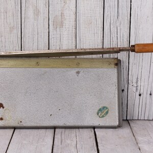 May include: Vintage paper cutter with a silver metal base and a wooden handle. The blade is extended, ready for use. The cutter has a measurement guide along the side and a small sticker on the front. The wooden handle is approximately 4 inches long.