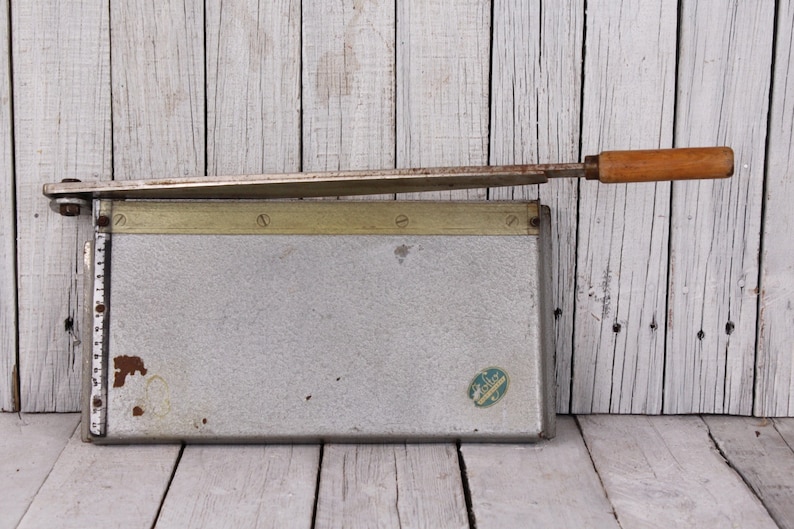 May include: Vintage paper cutter with a silver metal base and a wooden handle. The blade is extended, ready for use. The cutter has a measurement guide along the side and a small sticker on the front. The wooden handle is approximately 4 inches long.