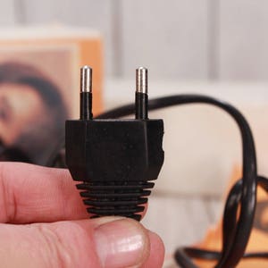 May include: A black electrical plug with two metal prongs. The plug is held in a hand, with a black cord extending from it. The background is blurred, showing a light-colored surface and a box.